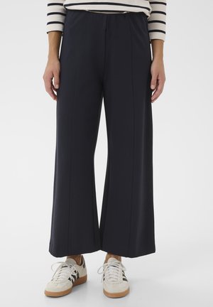 Personne portant un pantalon noir large et court, des baskets blanches avec des rayures noires, et une chemise blanche à manches longues avec des rayures horizontales noires.