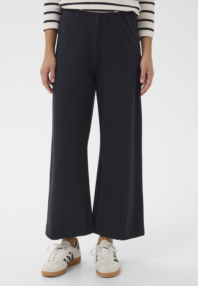 Personne portant un pantalon noir large et court, des baskets blanches avec des rayures noires, et une chemise blanche à manches longues avec des rayures horizontales noires.