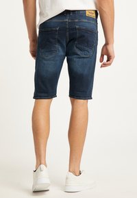 Pantalones cortos de mezclilla azul oscuro con un aspecto ligeramente desgastado, terminados con un dobladillo enrollado, bolsillos traseros con costura curva y una etiqueta de cuero.