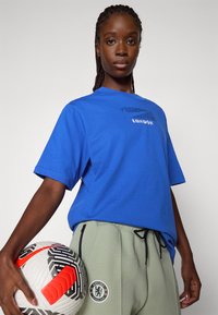 Blå bomullströja med ett tryckt grafik som läser "LONDON", i kombination med ljusgröna joggers som har en Chelsea FC-logotyp och svarta accenter. Håller i en fotboll.
