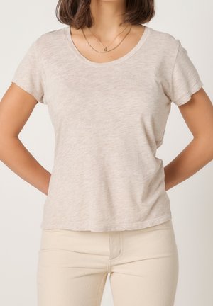 Vrouw met een beige T-shirt met korte mouwen en lichtgekleurde broek, handen achter haar rug, met gelaagde gouden kettingen.