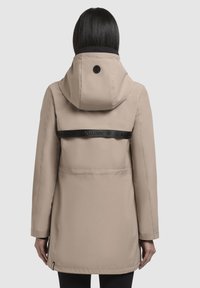 Beige vandtæt jakke med hætte, der har en sort rygrem, sømdetaljer og et logo ved halsen. Glat, let tekstur.