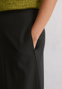 Schwarze Hosen mit glatter Textur, ausgestattet mit Seitentaschen, einem maßgeschneiderten Schnitt und einer sichtbaren Naht entlang des Beins. Kombiniert mit einem grünen Strickoberteil.