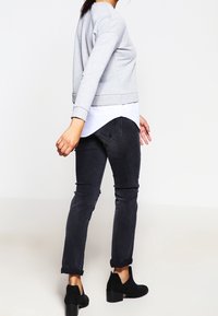 Mujer alejándose, vestida con un sudadera gris sobre una camisa blanca, jeans negros arremangados y botas de tacón negras hasta el tobillo, sobre un fondo blanco.