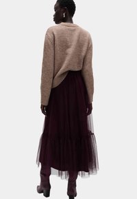 Kobieta stojąca tyłem, ubrana w beżowy sweter, ciemnofioletową spódnicę midi z tiulu, dopasowane botki oraz złote kolczyki w kształcie koła, na białym tle.