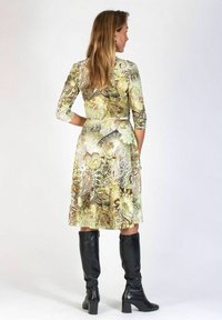 Nainen seisoo selkä kameraan päin, pukeutuneena polvimittaisiin vihreisiin ja ruskeisiin paisley-kuosisempiin mekkoihin, joissa on kolmanneksen pituiset hihat, sekä mustiin polvisukkiin. 