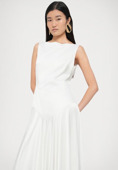 Balta, be rankovių suknelė su asimetrišku dizainu ir laisvai krentančia sijonu. Lygus audinio tekstūra, minimalistinis stilius, dėvima modelio su auksinėmis auskarais.