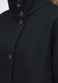 Manteau en laine noire avec col montant et quatre boutons marron fermés, montrant le haut du torse et une partie des épaules.