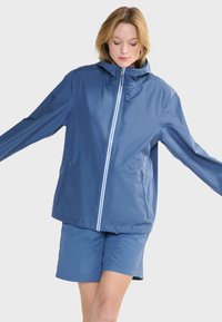 Giacca impermeabile blu chiaro con cappuccio, cerniera frontale e due tasche laterali. Abbinata a pantaloni corti coordinati, caratterizzati da una texture liscia.