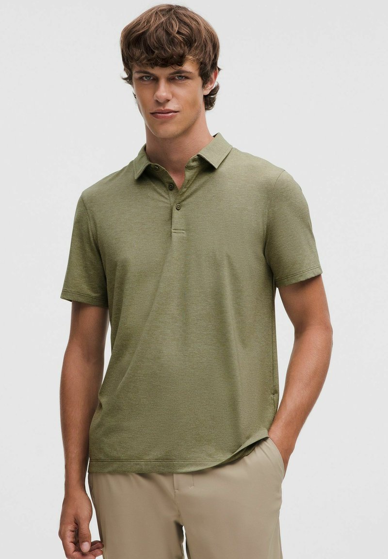 Oliwkowa koszulka polo z krótkim rękawem, zapinana na guziki, o miękkiej fakturze. Stylizowana z kremowymi spodniami, charakteryzuje się luźnym krojem.