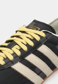 Black and gray sneaker with three white stripes, textured fabric, and yellow laces. Features a rubber sole and a gold logo detail.