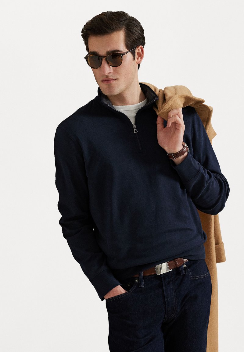 Mann iført mørke solbriller, marineblå quarter-zip genser og mørke jeans, holder en kamelfarget frakk over skulderen.