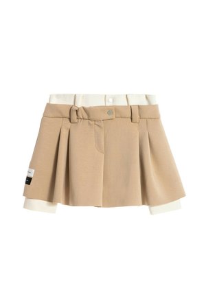 Minifalda plisada beige con pantalones cortos interiores crema incorporados, trabillas para cinturón, cierre de botón a presión y un pequeño parche negro y blanco en el lado izquierdo.