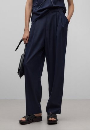 Frau trägt lockere dunkelblaue Hose, schwarze Sandalen mit offenem Zehenbereich und Absatz, hält eine kleine schwarze Handtasche, steht mit einer Hand in der Tasche.