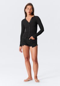 Černý dlouhý rukáv s knoflíkovou přední částí, spojený s roztrhanými černými džínovými šortkami. Ležérní žabky doplňují vzhled.