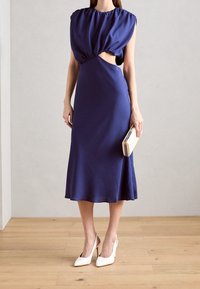 Marinblå satin klänning med puffärmar, utskärning, flödande kjol och knälång nederdel. Kombinerad med vita stilettklackar och en kuvertväska.