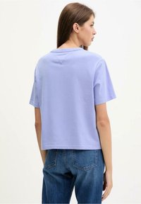 Levandulové tričko s krátkým rukávem, volným střihem, kulatým výstřihem a švem na zádech, doplněné modrými džínami.
