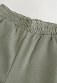 Pantaloni felpa verde chiaro con vita elastica e tessuto morbido e texturizzato. Le pieghe aggiungono dettagli al design.