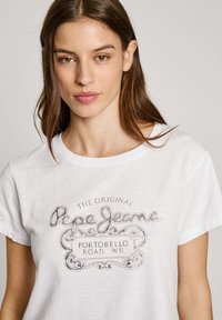 Balta medvilnės marškinėliai su pilku senoviniu logotipu ir dekoro elementais, kuriuose parašyta "Pepe Jeans Portobello Road W.11." Trumpomis rankovėmis.
