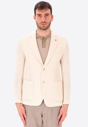 Uomo che indossa una giacca color crema con texture sopra una polo beige e pantaloni marrone chiaro, in piedi su sfondo uniforme.