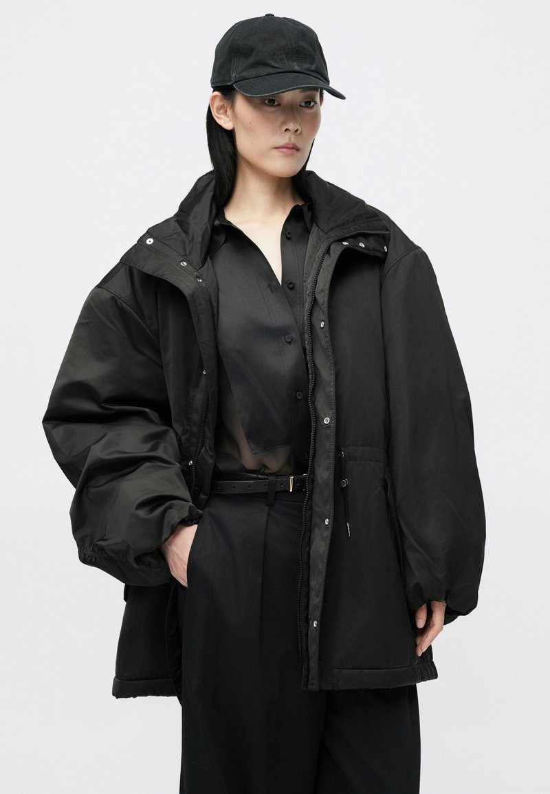 Black, oversized padded jacket with a high collar, zip closure, and snap buttons; paired with a sheer black shirt and black pants.