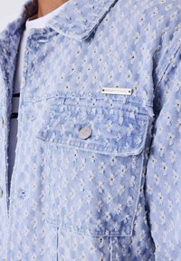 Lichtblauw spijkerjack met textuur en verweerde patronen, een enkele borstzak met een zilveren knoop en een merklabel op de kraag.