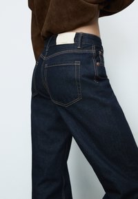 Tummansiniset farkut suoran leikkauksen kanssa, oransseilla ompeleilla, nahkalaminaatti, jossa lukee "D92", sekä kaksi läpällistä taskua takana.