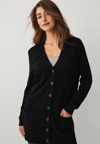 Zwarte cardigan met een v-hals, lange mouwen, knopen aan de voorkant, twee zakken en rib gebreide manchetten. Gemaakt van zachte, lichte stof.