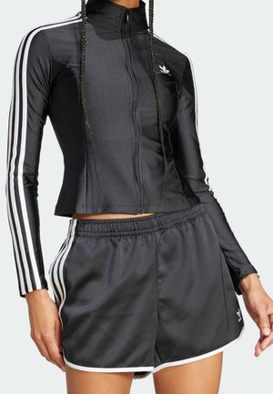 Femme portant une veste Adidas zippée noire et un short assorti avec des bandes blanches sur les manches et les côtés, debout avec une main légèrement levée.