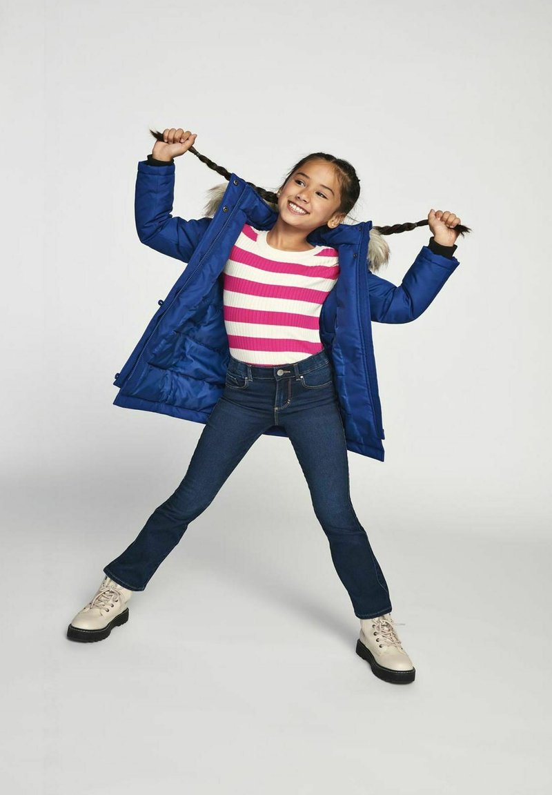 Parka blu isolato con cappuccio foderato in pelliccia sopra una maglietta a maniche lunghe rosa e bianca a righe, abbinata a jeans svasati e stivali beige.