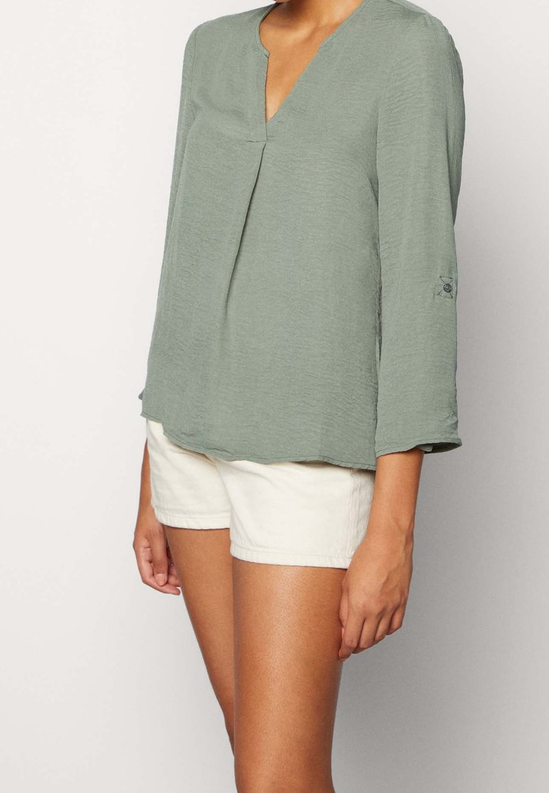 Blouse verte légère avec un col en V et des manches trois-quarts, présentant un détail de pli à l'avant et des poignets retroussés. Associée à un short clair.
