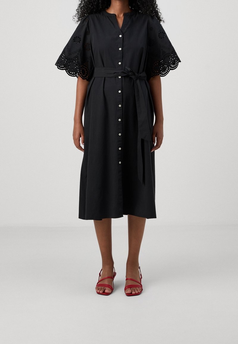 Czarna sukienka koszulowa z paskiem, z krótkimi haftowanymi rękawami i długością midi. W połączeniu z czerwonymi sandałami na paskach.