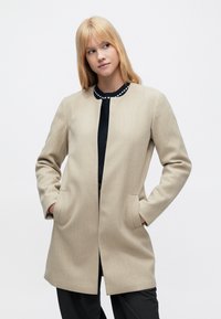 Μπεζ μάλλινο παλτό με ίσια γραμμή, μακριά μανίκια, δύο εμπρόσθιες τσέπες και στρογγυλή μαύρη λαιμόκοψη με λευκές λεπτομέρειες.