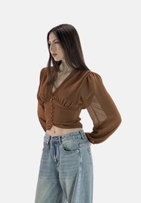 Blusa trasparente marrone con maniche a sbuffo, scollatura a V profonda, chiusura frontale con bottoni, abbinata a jeans azzurri a vita alta.