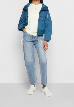 Persoon draagt een blauwe capuchonjas, een crèmekleurig T-shirt met opdruk, lichtblauwe rechte spijkerbroek en witte sneakers, staand tegen een effen achtergrond.