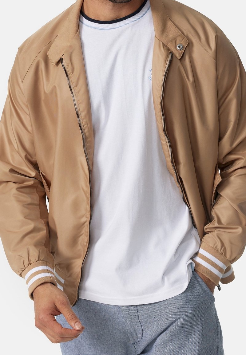 Homme portant une veste zippée beige avec des poignets rayés sur un t-shirt blanc et un pantalon bleu clair, une main dans la poche, cadré au niveau du cou.