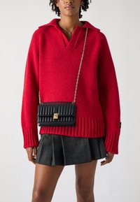 Red knitted sweater with a collar, paired with a black quilted crossbody bag featuring a gold accent. Gray plaid pleated skirt completes the outfit.