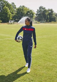 Adidas sportswear in blue with red accents, fitted design, long sleeves, and white stripes. Holding a black and white soccer ball on grass.