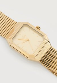 Ρολόι χειρός με ορθογώνιο χρυσό καντράν, χρυσή πλέξη μπρασελέ, μίνιμαλ σήματα στο καντράν, τρεις δείκτες και λογότυπο Lacoste στο μπρασελέ.