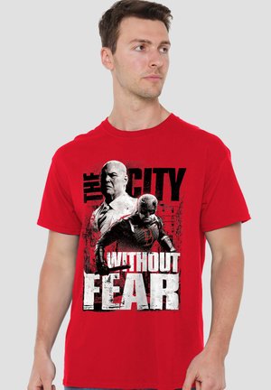 Man wearing a red t-shirt with graphic of two men, one in a suit and one in superhero costume, text "The City Without Fear."