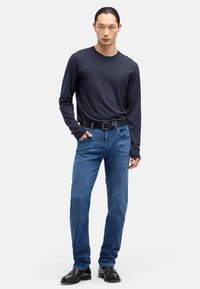 Niebieskie dżinsy denimowe o klasycznym kroju, zestawione z granatową koszulą z długim rękawem i czarnym paskiem. Model stoi w eleganckich butach na neutralnym tle.