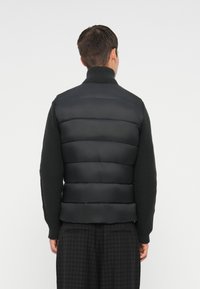 Gilet en duvet noir avec un col haut, un design matelassé et une texture lisse, associé à un pull en maille sombre en dessous.