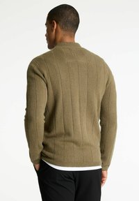 Bărbat îmbrăcat cu un pulover verde oliv cu un model în rigle și pantaloni negri, cu fața întoarsă și mâinile în buzunare, pe un fundal alb simplu.