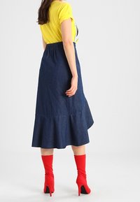 Gonna di jeans con orlo svasato e vita elasticizzata, abbinata a una camicia gialla a maniche corte e stivali rossi con tacco alto.