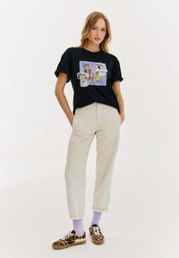 Czarna koszulka z graficznym nadrukiem, beżowe spodnie i trampki w panterkę. Posiada wywinięte mankiety i lawendowe skarpetki.