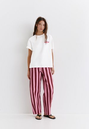 Jeune femme debout contre un mur blanc, portant une chemise blanche à manches courtes, un pantalon à rayures verticales rose et rouge foncé, et des sandales noires.