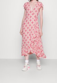 Florales Wickelkleid in Hellrosa mit roten Mustern, Flügelärmel und einem gerafften Saum. Kombiniert mit weißen Socken und hellen Sneakers.