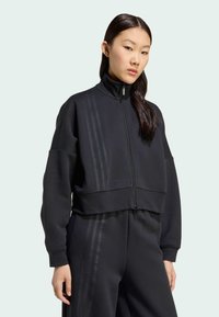Zwarte cropped ritsjack met hoge kraag, voorzien van verticale strepen en een losse pasvorm. De stof lijkt zacht met een gestructureerde afwerking.
