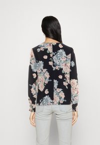 Blusa a maniche lunghe floreale realizzata in tessuto leggero. Presenta uno sfondo scuro con fiori pastello, maniche arricciate e un dettaglio con spacco sul retro.