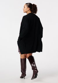 Vrouw met krullend haar in een paardenstaart, draagt grote oorbellen, een zwarte oversized cardigan, een jurk en kniehoge bruin leren laarzen met hak.